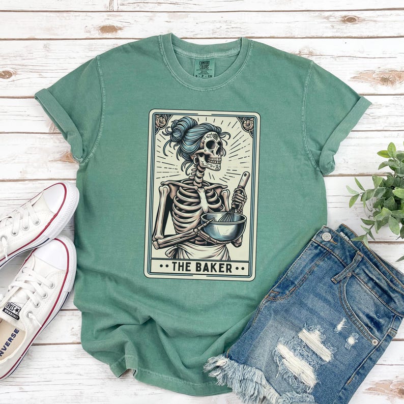 May include: A green t-shirt with a vintage-style illustration of a skeleton holding a bowl and whisk. The illustration is in a card-like frame with the text "The Baker" below it.