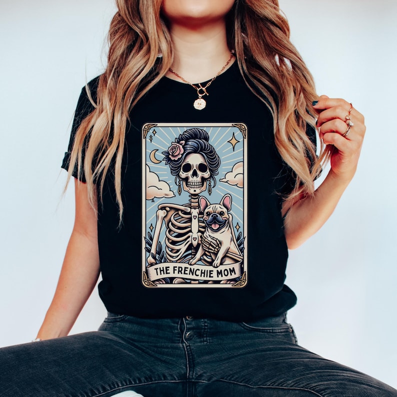 May include: Black t-shirt featuring a tarot card design with a skeleton woman holding a French Bulldog. The card has a light blue background with clouds and the text "THE FRENCHIE MOM". The skeleton has a floral headpiece.