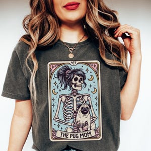 Puede incluir: Una camiseta gris oscuro con un gr&aacute;fico de un esqueleto sosteniendo un pug. El gr&aacute;fico est&aacute; en un estilo de carta de tarot con el texto "The Pug Mom" en la parte inferior.