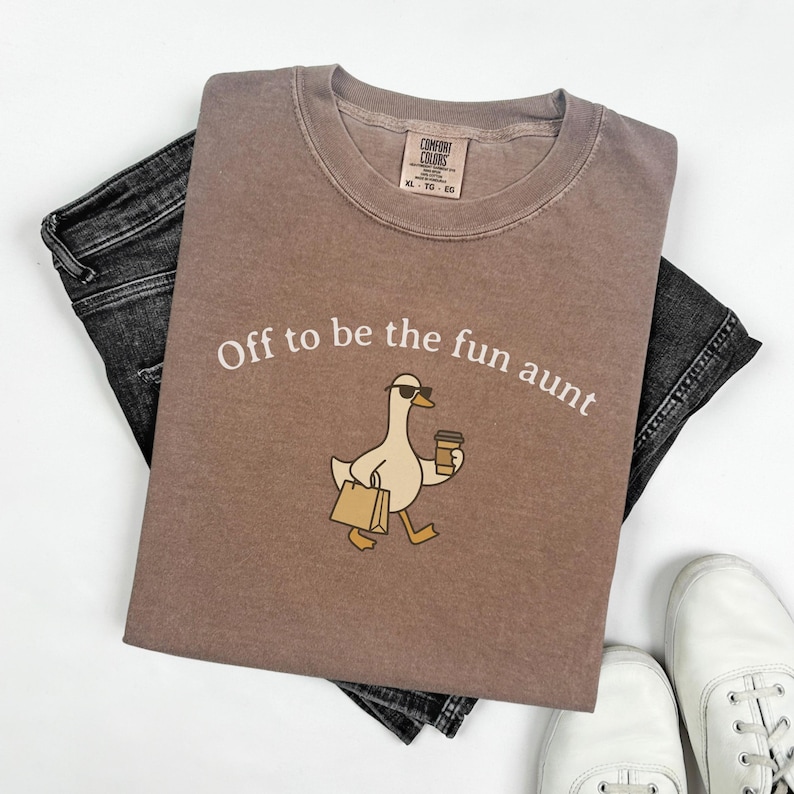 May include: A brown Comfort Colors t-shirt with the phrase "Off to be the fun aunt" and a cartoon duck wearing sunglasses and carrying a coffee cup and shopping bag. The shirt is laid on top of black denim jeans and white sneakers.