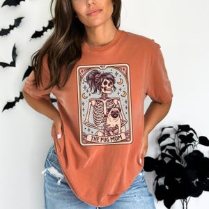 Puede incluir: Una camiseta naranja quemada con un gr&aacute;fico de un esqueleto sosteniendo un pug. El gr&aacute;fico est&aacute; en un estilo de carta de tarot con el texto "The Pug Mom" en la parte inferior.