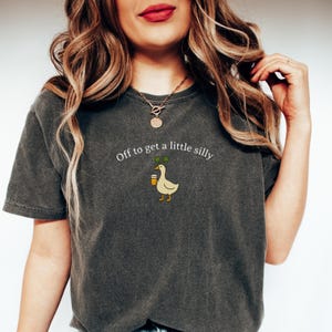 May include: Dark gray t-shirt with the phrase "Off to get a little silly" and a cartoon duck holding a drink. The duck has a sprig of green on its head. The shirt is a relaxed fit.