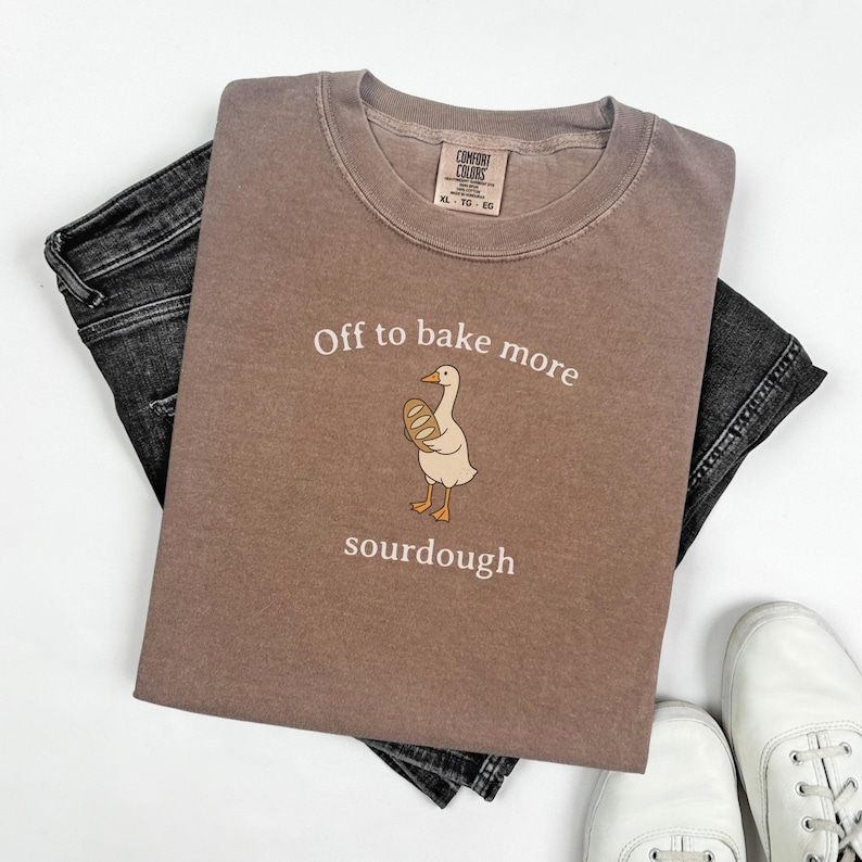 K&ouml;nnte beinhalten: Braunes Comfort Colors T-Shirt mit dem Text "Off to bake more sourdough" und einer Cartoon-Gans, die Brot h&auml;lt. Das Shirt liegt auf einer schwarzen Jeans und wei&szlig;en Turnschuhen.