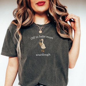 K&ouml;nnte beinhalten: Dunkelgraues T-Shirt mit dem Text "Off to bake more sourdough" und einer Cartoon-Gans, die Brot h&auml;lt. Das T-Shirt hat einen l&auml;ssigen Stil.