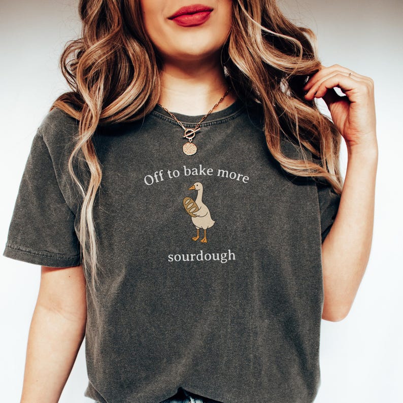 K&ouml;nnte beinhalten: Dunkelgraues T-Shirt mit dem Text "Off to bake more sourdough" und einer Cartoon-Gans, die Brot h&auml;lt. Das T-Shirt hat einen l&auml;ssigen Stil.