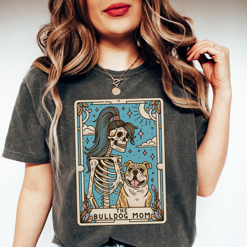 May include: A grey t-shirt featuring a graphic of a skeleton with a ponytail holding a bulldog. The graphic is in the style of a tarot card and reads "The Bulldog Mom" at the bottom.