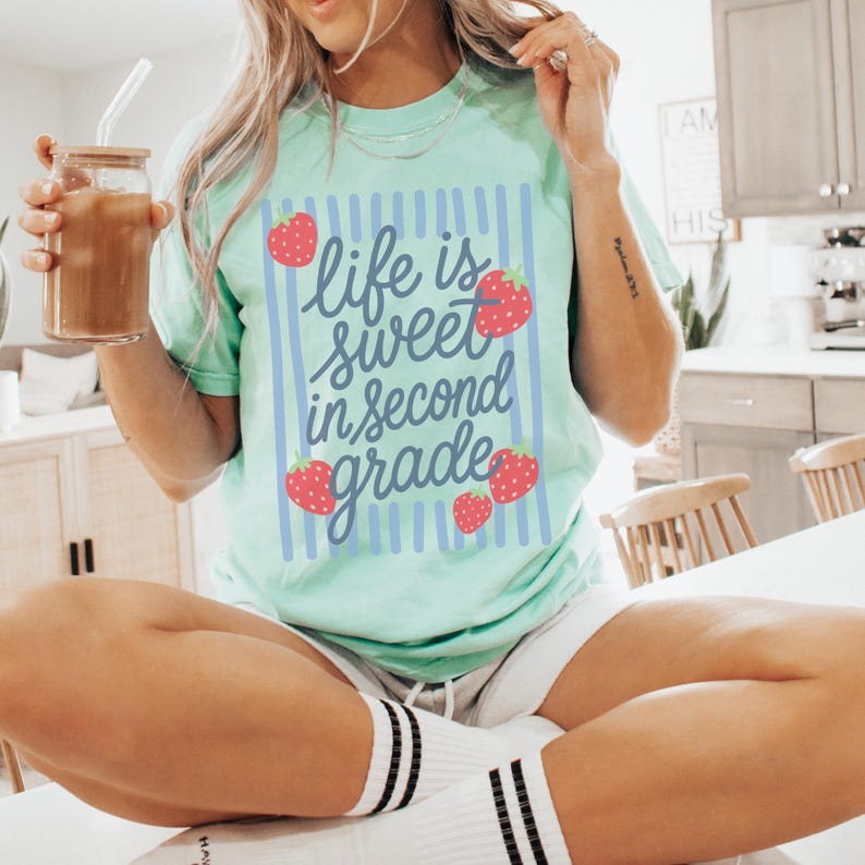 K&ouml;nnte beinhalten: Hellgr&uuml;nes T-Shirt mit dem Schriftzug "life is sweet in second grade" in blauer Schrift, verziert mit roten Erdbeeren. Das T-Shirt hat blaue vertikale Streifen und einen l&auml;ssigen Stil.