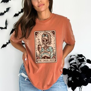 Pu&ograve; includere: Una maglietta color arancio bruciato con una grafica di uno scheletro che indossa un'uniforme da cheerleader e tiene una tazza di caff&egrave;. La grafica &egrave; in stile carta dei tarocchi con il testo "The Cheer Mom".