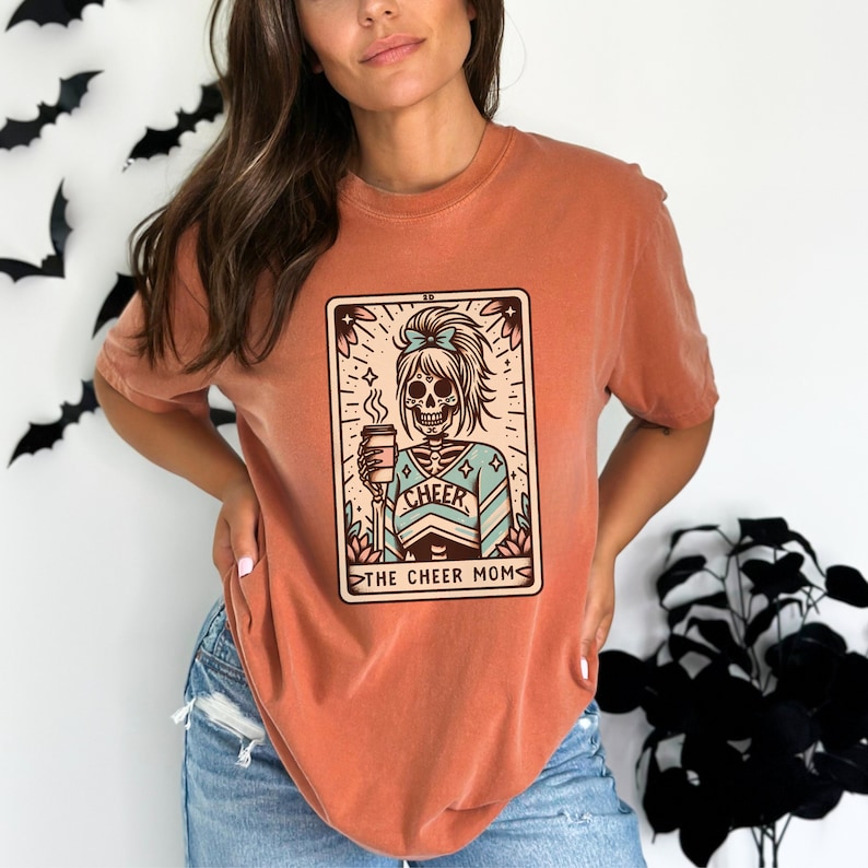 Pu&ograve; includere: Una maglietta color arancio bruciato con una grafica di uno scheletro che indossa un'uniforme da cheerleader e tiene una tazza di caff&egrave;. La grafica &egrave; in stile carta dei tarocchi con il testo "The Cheer Mom".
