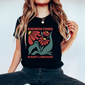 May include: Black t-shirt with a graphic of red flowers and green leaves. The text "Kindness comes in many languages" is printed on the shirt.