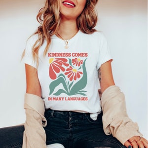 May include: White t-shirt with a graphic of red and yellow flowers and green leaves. The text "Kindness comes in many languages" is printed on the shirt.