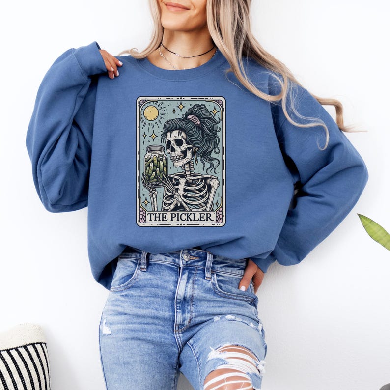 May include: A blue sweatshirt featuring a tarot card design with a skeleton holding a jar of pickles. The card has a sun and stars design, with the text "THE PICKLER" at the bottom. The sweatshirt is a solid blue color.