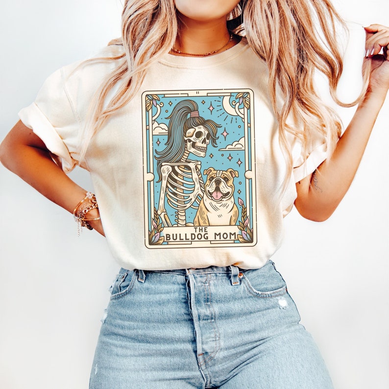May include: A white t-shirt featuring a graphic of a skeleton holding a bulldog. The graphic is in a tarot card style with the text "The Bulldog Mom".