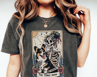 May include: Dark grey t-shirt featuring a graphic of a skeleton with a corgi dog. The skeleton has a floral hairpiece and is embracing the dog. The text "THE CORGI MOM" is displayed on a banner below the image. The design is set within a rectangular frame.