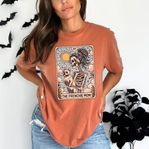 May include: A burnt orange t-shirt with a graphic of a skeleton holding a french bulldog. The graphic is in the style of a tarot card and says "The Frenchie Mom" at the bottom.