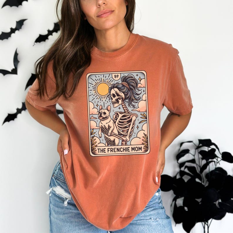 May include: A burnt orange t-shirt with a graphic of a skeleton holding a french bulldog. The graphic is in the style of a tarot card and says "The Frenchie Mom" at the bottom.