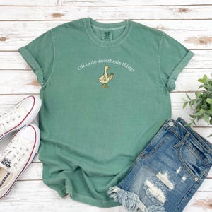 May include: A sage green t-shirt with rolled sleeves displaying the text "Off to do anesthesia things" and a cartoon goose with a coffee cup. The shirt is styled with denim shorts and white trainers.