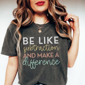 May include: Dark gray t-shirt with the text "BE LIKE subtraction AND MAKE A difference" in white, yellow, pink, and teal lettering. The shirt has a vintage, worn look.