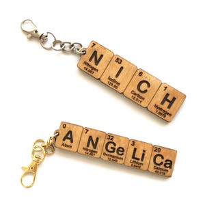 Peut inclure: Deux porte-clés en bois avec les noms "NICH" et "ANGE Li Ca" épelés à l'aide d'éléments du tableau périodique. Chaque porte-clés a un fermoir et une chaîne en métal. Le porte-clés "NICH" est en haut, et le porte-clés "ANGE Li Ca" est en bas.