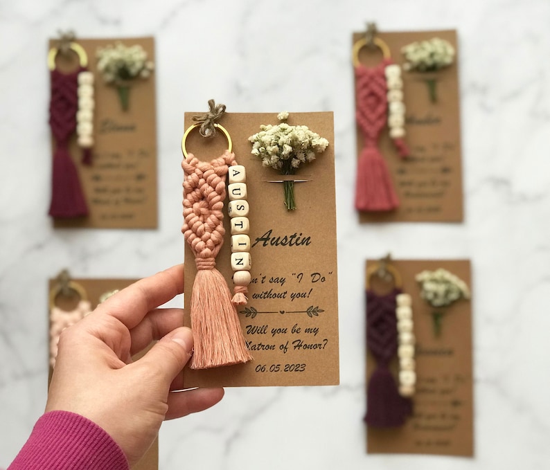 May include: A peach-colored macrame keychain with a wooden bead charm that spells "Austin". The keychain is attached to a card with the text "Can't say "I Do" without you! Will you be my Matron of Honor? 06.05.2023"