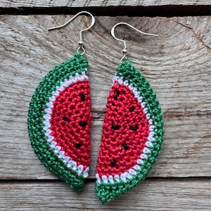 May include: A pair of handmade crochet earrings shaped like watermelon slices. The earrings feature a red center with black seed details, a white rind, and a green outer edge. The earrings hang from silver-colored fish hook ear wires.