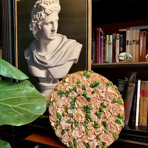 May include: A round, decorative wall art piece featuring a relief of roses in shades of peach and green. The artwork is displayed in a room with a bust of a classical figure and a framed portrait. A bookshelf filled with books is in the background.