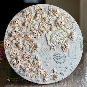 May include: Circular canvas art with a 3D floral design. It features cream and beige roses, berries, and leaves. The background has a vintage print with stamps and script. This decorative artwork would suit any home.