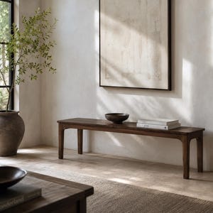 May include: A dark brown wooden bench with a rectangular top and four legs. A dark bowl and two books are on the bench. The bench is in a room with a large window, a large vase with a plant, and a framed piece of art.