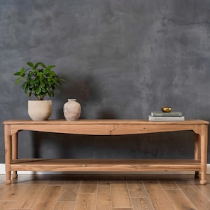 Puede incluir: Una consola de madera marrón claro con un estante inferior y patas curvas. Una planta en maceta, un jarrón y libros con un cuenco dorado se exhiben sobre la mesa. La mesa está colocada contra una pared gris oscuro.