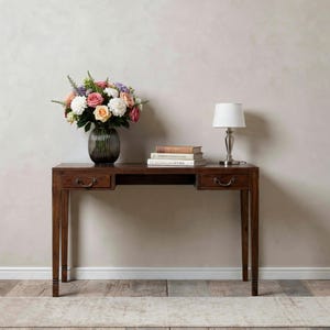 May include: A dark brown wooden desk with two drawers, a vase of flowers, a lamp, and books. The flowers are in a clear glass vase. The lamp has a white shade and a silver base. The desk is on a light-coloured rug.