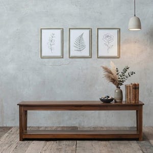May include: A long wooden bench with a lower shelf, topped with a ceramic vase containing dried foliage, a small dark bowl with stones, and three vintage books. Three framed line art prints of a eucalyptus branch, a fern, and a rose hang above.