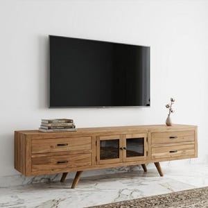 May include: A light brown wooden TV stand with two drawers on each side and glass-fronted cabinets in the center. The stand has angled legs and a stack of books on top. A black flat-screen TV is mounted on the white wall above.