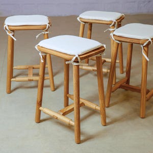 Puede incluir: Cuatro taburetes de bar de madera con marcos de madera de color claro y asientos acolchados blancos. Los asientos son cuadrados y están sujetos con lazos blancos. Los taburetes tienen un diseño simple y minimalista, con reposapiés y patas resistentes.