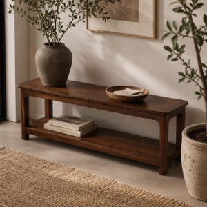 May include: A dark brown wooden bench with a lower shelf, holding two books. A large, textured vase with greenery sits on the bench, along with a wooden bowl. A potted plant is to the right, and a woven rug is in the foreground.