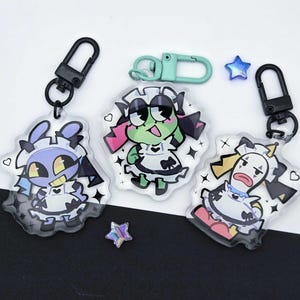 May include: Three acrylic keychains featuring cartoon characters in maid outfits with black clasps, including a light blue character, a green character with a mint clasp, and a white character, accompanied by two small blue and purple star charms.