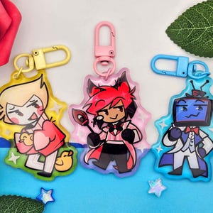May include: Three cartoon character keychains with colorful designs. One keychain has a yellow clip and a character in a red coat. The second keychain has a pink clip and a character in a pink coat. The third keychain has a blue clip and a character in a white suit.