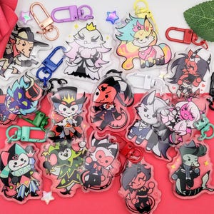 May include: A collection of clear acrylic keychains featuring colorful cartoon characters with various designs. Each keychain has a metal clasp in different colors, including gold, purple, blue, and red. The keychains are arranged on a red surface with decorative elements.
