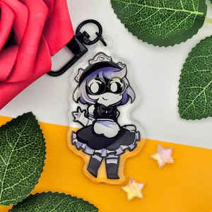 May include: A clear acrylic keychain featuring a cartoon maid character with purple hair, wearing a black and white maid outfit. The keychain has a black clasp and is surrounded by decorative elements.