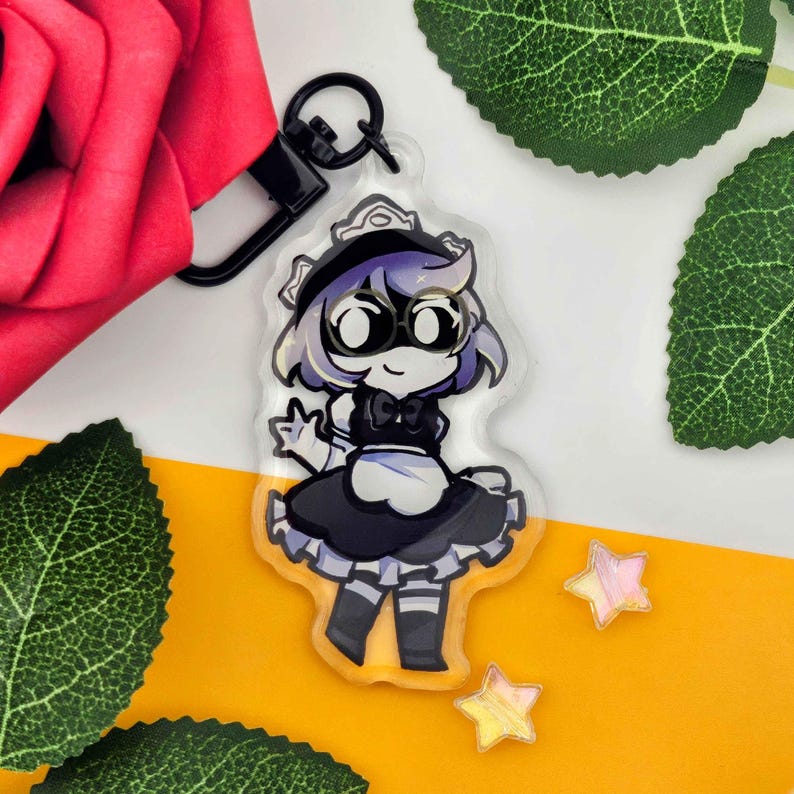 May include: A clear acrylic keychain featuring a cartoon maid character with purple hair, wearing a black and white maid outfit. The keychain has a black clasp and is surrounded by decorative elements.