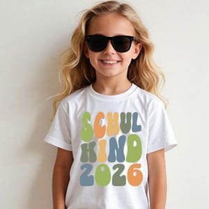 Könnte beinhalten: Ein weißes T-Shirt mit kurzen Ärmeln und dem Text SCHUL KIND 2026 in einer wellenförmigen, mehrfarbigen Schriftart. Die Buchstaben und Zahlen sind in den Farben Salbeigrün, gedämpftes Orange und Staubblau mittig auf dem T-Shirt aufgedruckt.