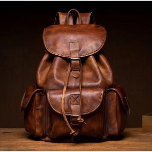 Pode incluir: Uma mochila de couro castanho com fecho de aba e dois bolsos frontais. A mochila tem alças ajustáveis e uma pega superior. O couro tem um aspeto envelhecido.