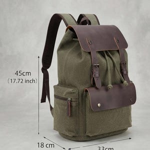 Puede incluir: Una mochila de lona verde oliva con detalles de cuero marrón oscuro, que cuenta con un cierre de cordón, dos correas con hebilla en la parte delantera y un bolsillo lateral con cremallera. La mochila mide 45 cm de alto, 33 cm de ancho y 18 cm de fondo.