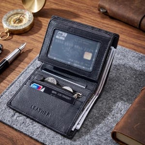 Op de afbeelding: Een zwarte bi-fold portemonnee met een textuur en het woord LEATHER in reliëf op de voorzijde. De portemonnee heeft een transparant ID-venster, meerdere kaartsleuven, een vak met ritssluiting voor munten en een vak voor papiergeld.
