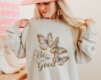 May include: A light beige sweatshirt featuring a butterfly graphic with the text "Be the Good". The butterflies are in shades of brown, orange, and teal. The sweatshirt is a relaxed fit.