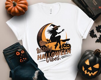 Puede incluir: Camiseta blanca con una silueta negra de una bruja volando en una escoba sobre una luna creciente. El texto "Witchy Halloween Vibes" está impreso en naranja y negro.