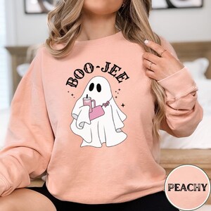 May include: A peach-colored sweatshirt with a ghost holding a pink cup and a pink purse. The ghost has a black outline and the text "BOO-JEE" is written above the ghost in black.