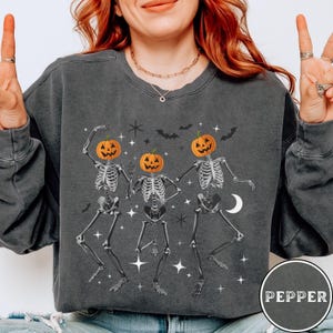 May include: A gray sweatshirt with a graphic of three skeletons wearing pumpkin heads and dancing. The skeletons are outlined in white and the pumpkins are orange with black stems. The sweatshirt has a distressed look and the text "PEPPER" is printed on the bottom right corner.