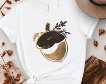 Puede incluir: Camiseta blanca con un diseño de bellota dorada y marrón. La bellota tiene un aspecto texturizado y está rodeada de hojas.