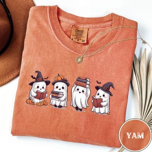 以下が含まれることがあります： ハロウィーンをテーマにしたデザインの、ヤム色のTシャツ。デザインには、本を持ったり、魔女の帽子やカボチャの帽子をかぶったりしている4匹の漫画の幽霊が描かれています。このシャツはクルーネックで、「Comfort Colors」と書かれた小さなタグが付いています。