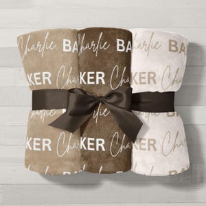 May include: Three plush blankets in shades of beige and brown, tied with a dark brown ribbon and bow. Each blanket is printed with the name "Charlie Baker" in a cursive font. The blankets are folded and stacked together.